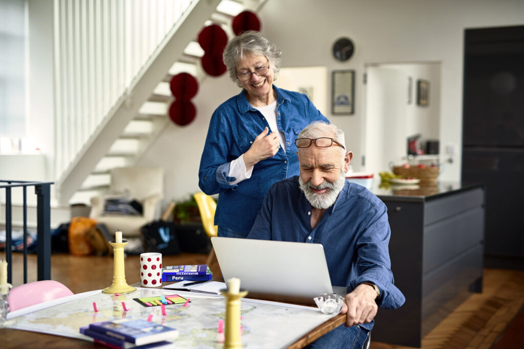 Oud stel kijkt lachend naar de laptop terwijl de man aan tafel zit en de vrouw achter hem staat met een bril op