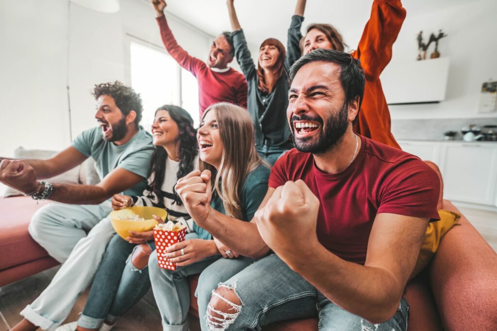 7 mensen kijken naar TV terwijl ze op de bank zitten en erachter staan en juichen